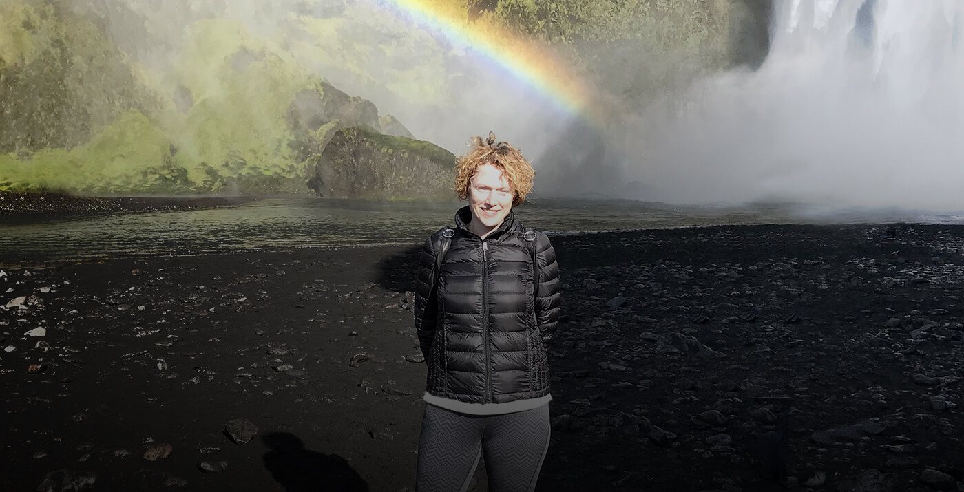 Maria Braun-Perez standing at dramatic Icelandic waterfall with rainbow through mist on black volcanic sand beach, showcasing adventure and creative spirit